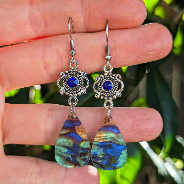 A pair of dangling earrings held in a person's hand, featuring ornate silver-toned filigree tops with deep blue round stones and teardrop-shaped iridescent blue-green gold-toned pendants, against a blurred green outdoor background.