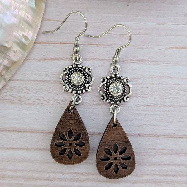 Pair of earrings with wooden teardrop pendants and silver floral designs on a light wood surface.