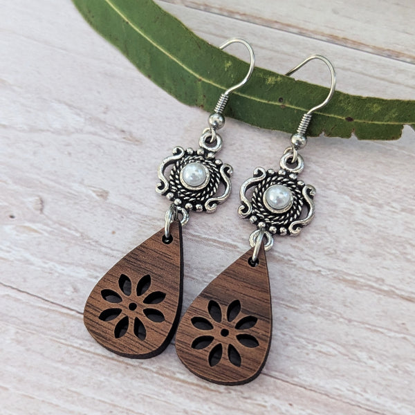 A pair of bohemian-style dangle earrings featuring carved dark brown wooden teardrop pendants with floral cutouts and ornate silver-toned metal tops with pearl-like centers, displayed on a light wood surface with a green leaf.