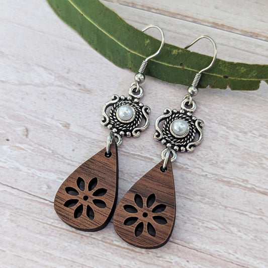 A pair of bohemian-style dangle earrings featuring carved dark brown wooden teardrop pendants with floral cutouts and ornate silver-toned metal tops with pearl-like centers, displayed on a light wood surface with a green leaf.