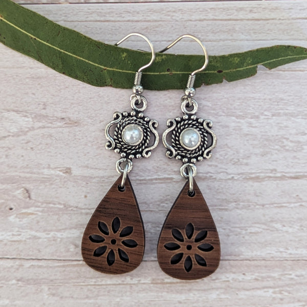 A pair of bohemian-style dangle earrings with silver filigree details, white pearl-like centers, and dark brown wooden teardrop charms with laser-cut floral patterns, displayed on a green leaf over a light wood background.