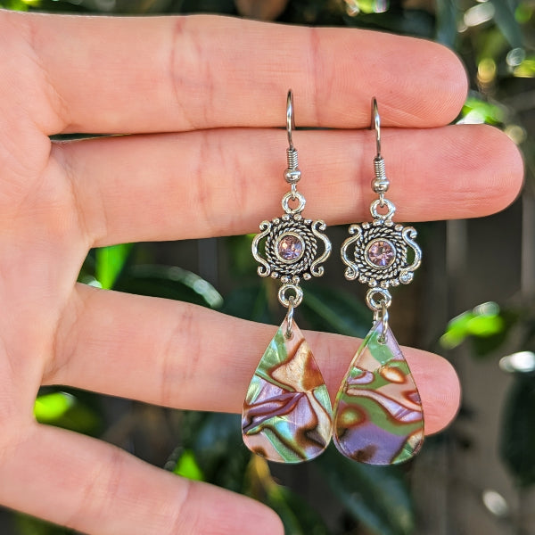 A pair of dangling earrings held on a person's hand, featuring silver-toned filigree tops with small pinkish-purple stones and teardrop-shaped acrylic drops with iridescent green, purple, and gold marbling, against a blurred green outdoor background.