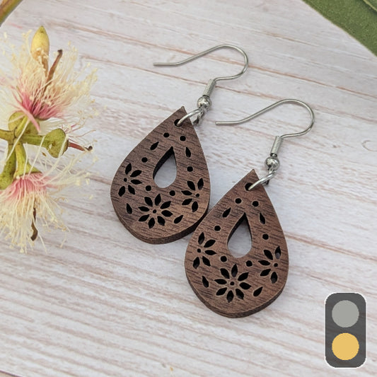 Wooden teardrop earrings with floral cutouts on a light wooden surface with flowers. Stainless steel hooks.