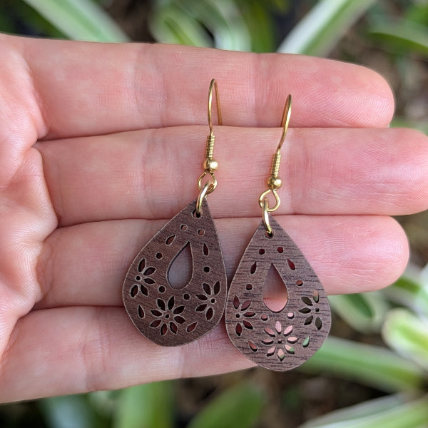 Wooden teardrop earrings with floral cutouts held between fingers against a blurred natural background. 18k gold plated stainless steel hooks.