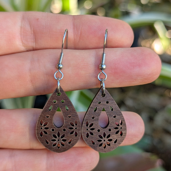 Wooden teardrop earrings with floral cutouts held between fingers against a blurred natural background. Stainless steel hooks.