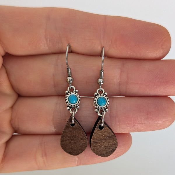 Pair of earrings with wooden teardrop pendants and turquoise stones held between fingers. Stainless steel hooks.