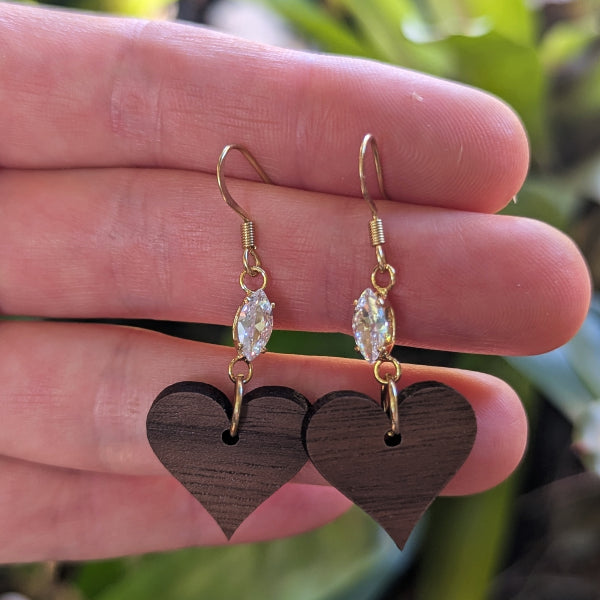 Gold wooden heart earrings with clear stones held between fingers against a blurred natural background