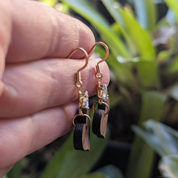 Gold wooden heart and crystal earrings held between fingers with a blurred green plant background. Side view.