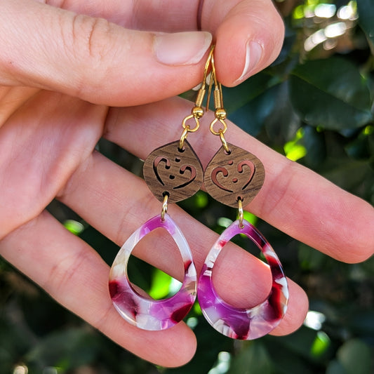 A close-up of a hand holding a pair of dangling earrings against a blurred background of green leaves. The earrings feature laser cut wooden heart-shaped dangles with gold hooks and teardrop-shaped resin pendants in a marbled pink, purple, and white pattern.
