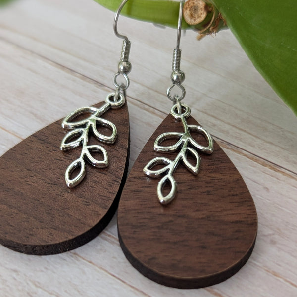 A pair of teardrop-shaped wooden drop earrings with a silver leaf branch charm, displayed on a light wood surface next to a green leaf.