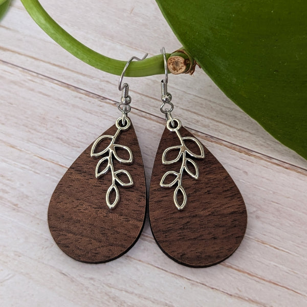 A pair of teardrop-shaped wooden drop earrings with a silver leaf branch charm, displayed on a light wood surface next to a green leaf.