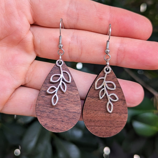 A pair of teardrop-shaped wooden dangle earrings with silver leaf filigree charms, held in a person's hand against a blurred green outdoor background.