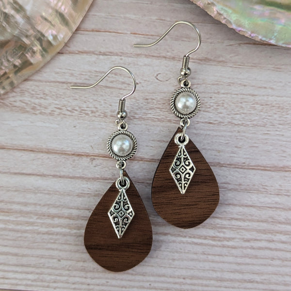 A pair of bohemian-style dangle earrings with dark brown teardrop wooden pendants, silver filigree diamond charms, and white faux pearl beads on silver hook wires, displayed on a light wood surface.