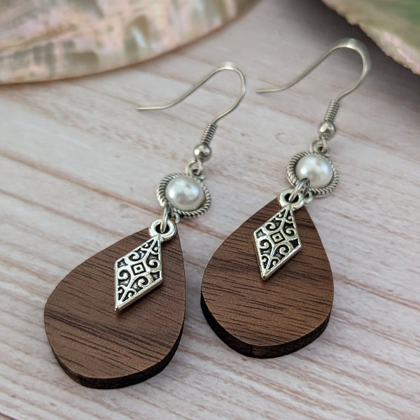 A pair of bohemian-style dangle earrings with dark brown teardrop wooden pendants, silver filigree diamond charms, and white faux pearl beads on silver hook wires, displayed on a light wood surface.