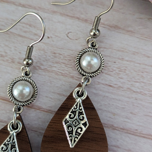 A pair of bohemian-style dangle earrings with dark brown teardrop wooden pendants, silver filigree diamond charms, and white faux pearl beads on silver hook wires, displayed on a light wood surface.