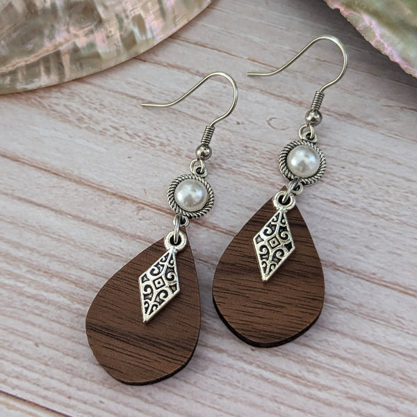 A pair of bohemian-style dangle earrings with dark brown teardrop wooden pendants, silver filigree diamond charms, and white faux pearl beads on silver hook wires, displayed on a light wood surface.