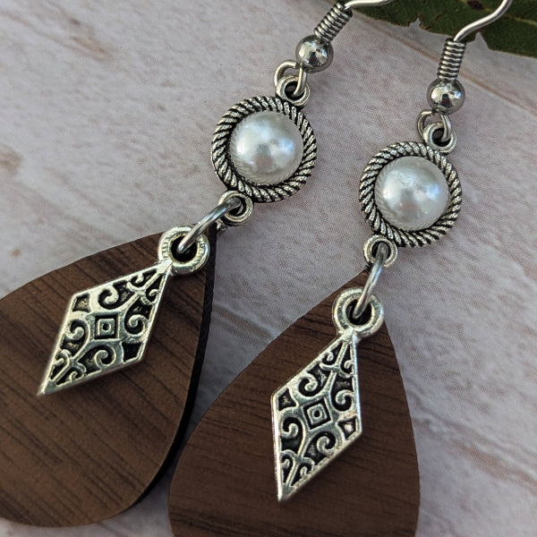 A pair of bohemian-style dangle earrings featuring oval dark brown wooden teardrop charms with ornate silver filigree cutouts, connected to round silver bezel settings holding white faux pearls, and hanging from silver fishhook wires.
