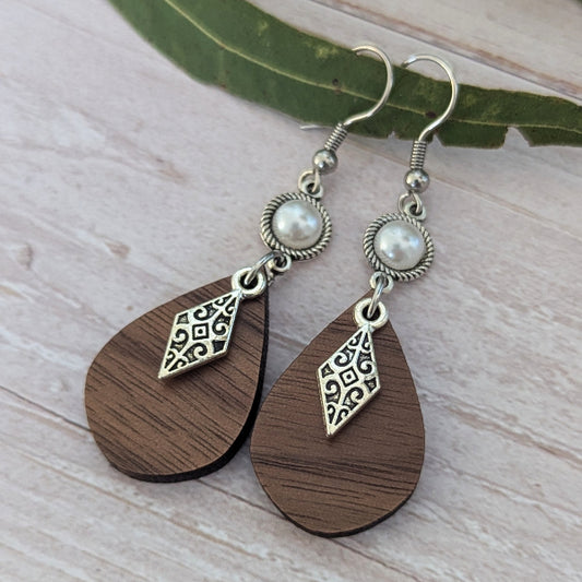 A pair of handmade dangle earrings with dark brown teardrop wooden pendants, silver filigree charms, faux pearl beads, and silver fishhook wires, displayed on a light wood surface with a green leaf.