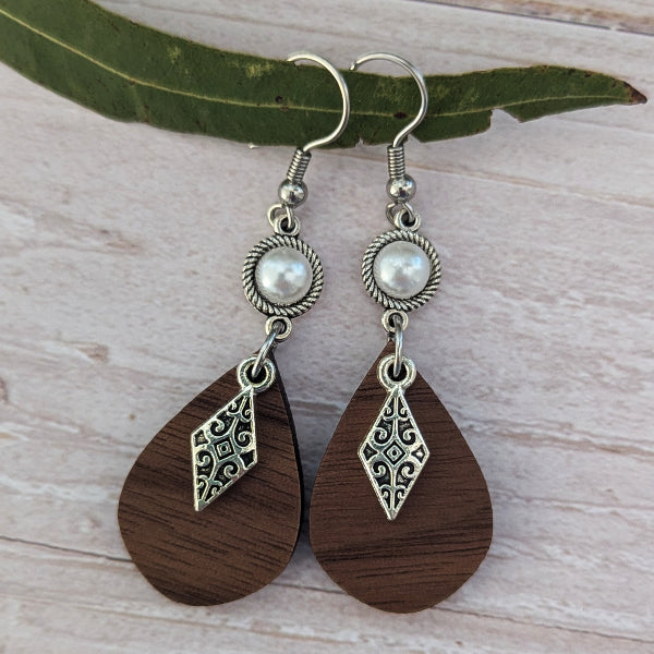 A pair of bohemian-style dangle earrings featuring dark brown teardrop wooden pendants with silver filigree details, a silver twisted bezel, and a white faux pearl, displayed on a light wood surface with a green leaf.