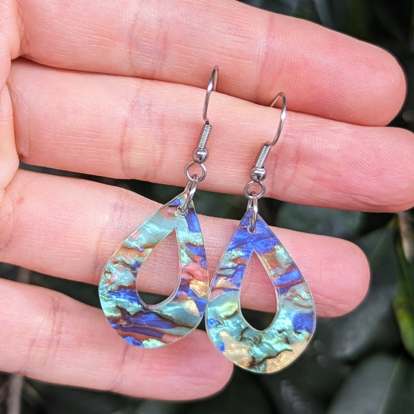A pair of teardrop-shaped dangling earrings with iridescent blue, green, and gold marbled inlay, held in a person's hand against a dark green leafy background.