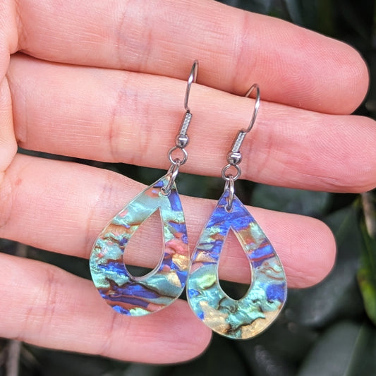 A pair of teardrop-shaped dangling earrings with iridescent blue, green, and gold marbled inlay, held in a person's hand against a dark green leafy background.