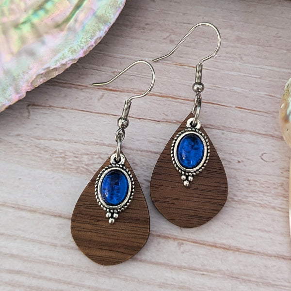 Wooden teardrop earrings with blue stones and silver accents on a wooden surface.
