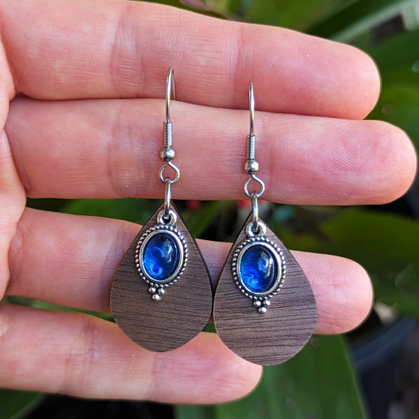 Wooden teardrop-shaped earrings with blue stones held between fingers against a blurred green background