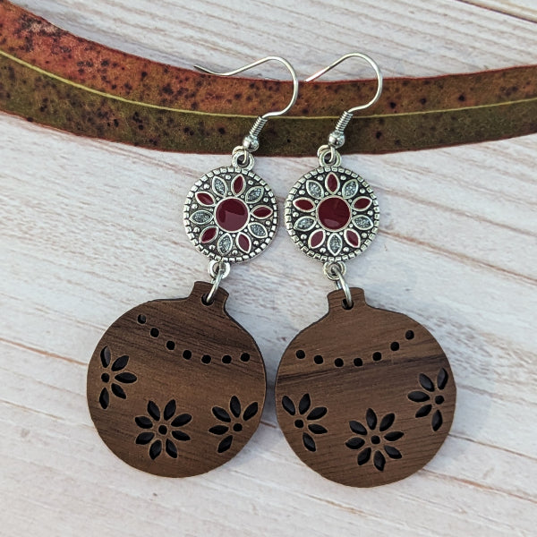 A pair of bohemian-style dangle earrings featuring oval brown wooden pendants with floral laser-cut patterns and circular silver-toned floral charms with red and silver accents, displayed on a light wood background with a dried leaf.