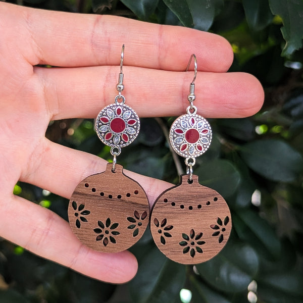 A pair of handmade dangle earrings held in a hand, featuring round wooden ornament-shaped drops with floral laser-cutout details and silver-toned floral connectors with red inlay, set against a background of green leaves.