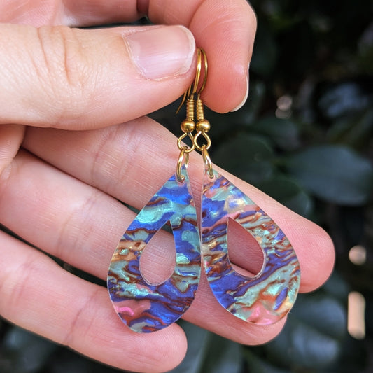 Teardrop-shaped earrings with iridescent acrylic inlays showing blue, purple, green, and pink tones, suspended from gold fishhook ear wires and held in a hand against a blurred dark green background.