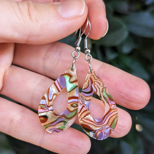 A close-up of a hand holding a pair of teardrop-shaped earrings with iridescent acrylic inlays and silver fishhook ear wires, set against a blurred green outdoor background.