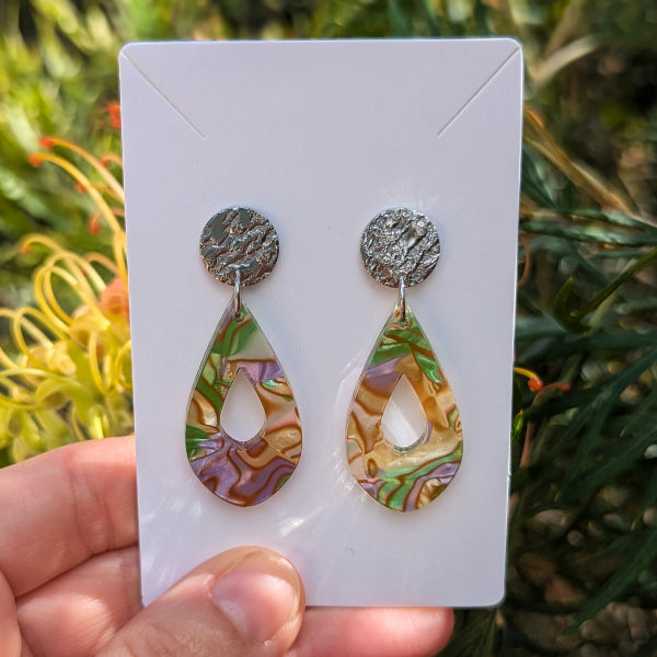 A pair of teardrop-shaped drop earrings with textured silver studs and iridescent multicolor acrylic drops, held on a white display card by a hand against a blurred green garden background.
