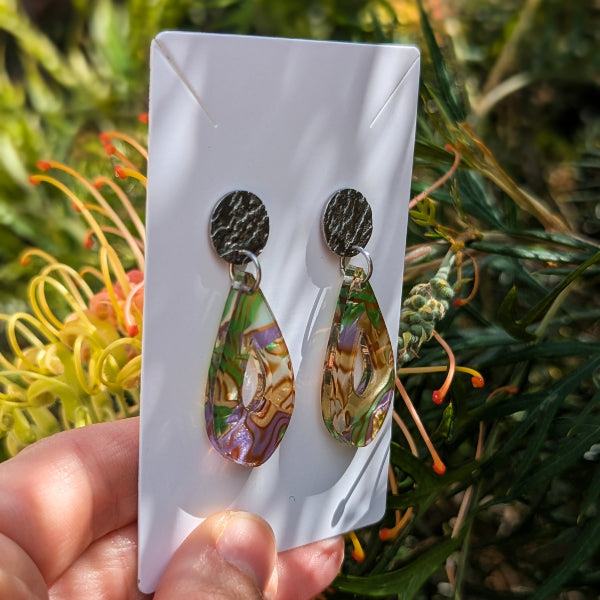 A pair of teardrop-shaped dangling earrings with textured dark metal tops and marbled green, gold, and purple acrylic drops, displayed on a white card held by a hand against a blurred outdoor garden background.