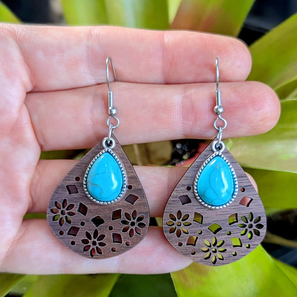 Wooden teardrop earrings with turquoise stones held in a hand against a green leafy background. Stainless steel hooks.