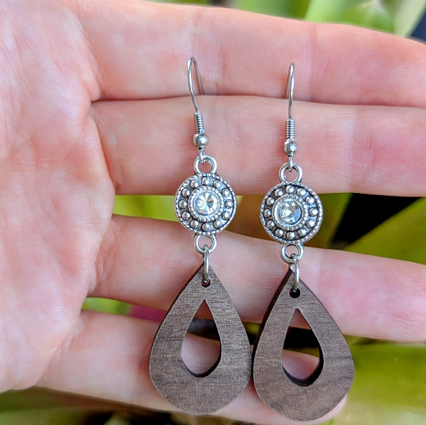 Wooden teardrop earrings with silver circular crystal accents held on a hand. Stainless steel hooks.