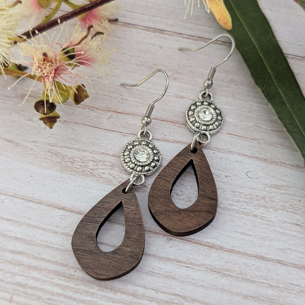 Wooden teardrop earrings with silver circular crystal accents on a light wooden surface with flowers in the background. Stainless steel hooks.