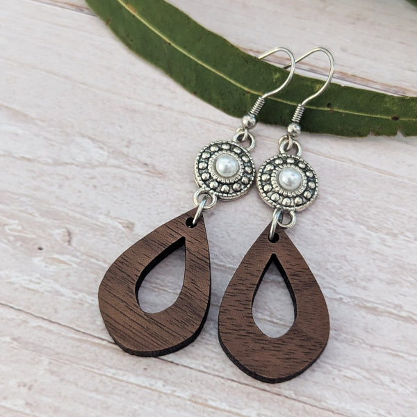 A pair of teardrop-shaped wooden dangle earrings with silver-toned metal hoops and circular silver-toned studs featuring white faux pearls, displayed on a light wood surface with a green leaf.