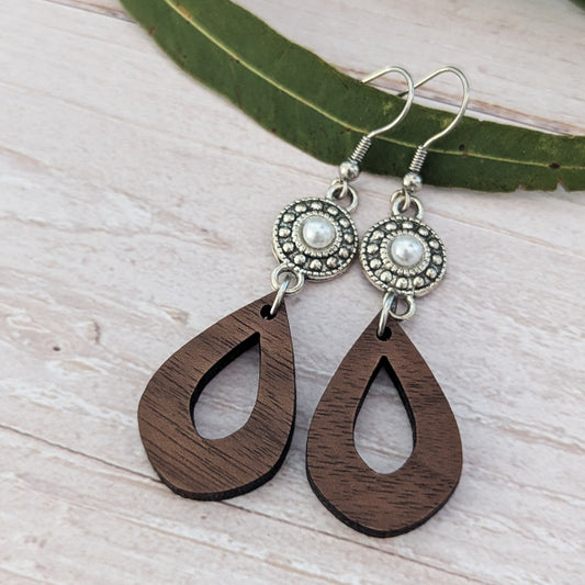 A pair of teardrop-shaped wooden dangle earrings with silver-toned metal hoops and circular silver-toned studs featuring white faux pearls, displayed on a light wood surface with a green leaf.