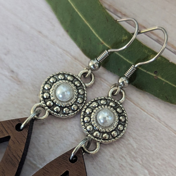 A pair of silver-toned dangle earrings with circular oxidized metal pendants featuring a central white faux pearl, displayed on a light wood surface with a green leaf and wooden props.