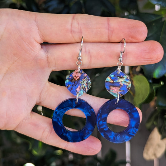 A pair of dangling earrings held in a hand, featuring large circular blue marbled acrylic drops and smaller iridescent blue top pieces with silver fishhook wires, against a blurred green foliage background.
