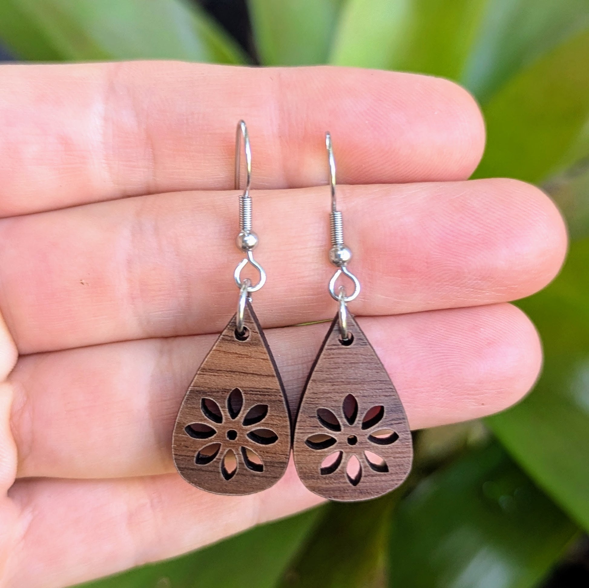 Wooden teardrop earrings with floral cutout held between fingers against a blurred green background. Stainless steel hooks.