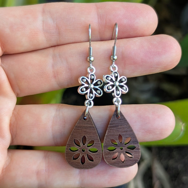 Wooden teardrop earrings with silver toned floral designs held between fingers. Stainless steel hooks.