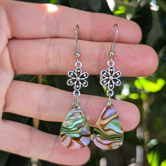A pair of dangling earrings held in a hand, featuring silver-tone metal flower charms and teardrop-shaped acrylic drops with iridescent green, pink, and gold swirls, against a blurred green foliage background.