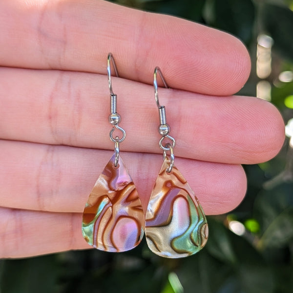 A pair of teardrop-shaped dangle earrings with iridescent acrylic drops showing pink, green, and gold tones, suspended from silver fishhook wires and held against a hand with a blurred green background.