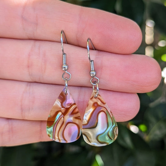 A pair of teardrop-shaped dangle earrings with iridescent acrylic drops showing pink, green, and gold tones, suspended from silver fishhook wires and held against a hand with a blurred green background.