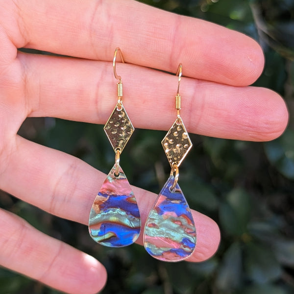 A pair of gold-tone dangle earrings with textured diamond-shaped tops and teardrop-shaped iridescent acrylic drops, held between fingers against a blurred green outdoor background.