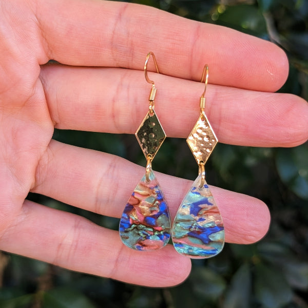 A pair of gold-tone dangle earrings with textured and polished diamond-shaped charms and teardrop-shaped acrylic drops, held in a person's hand against a blurred green background.