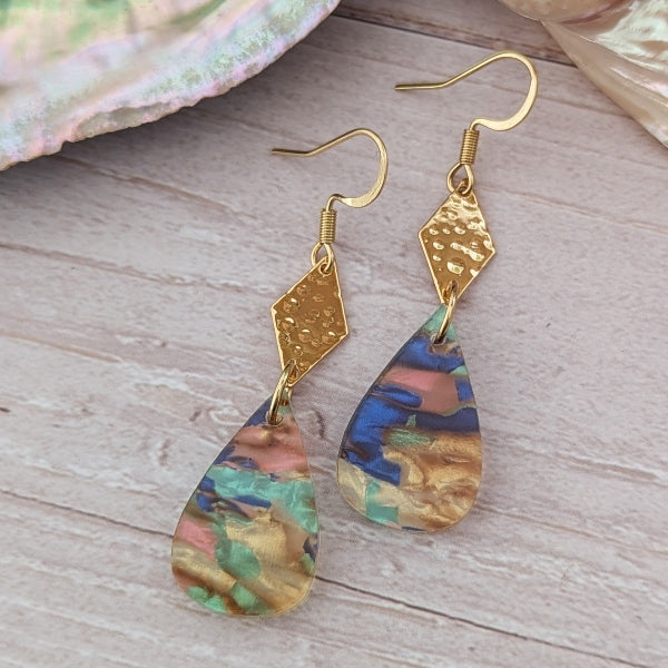 Gold earrings with teardrop-shaped, multicolored shapes on a wooden surface.
