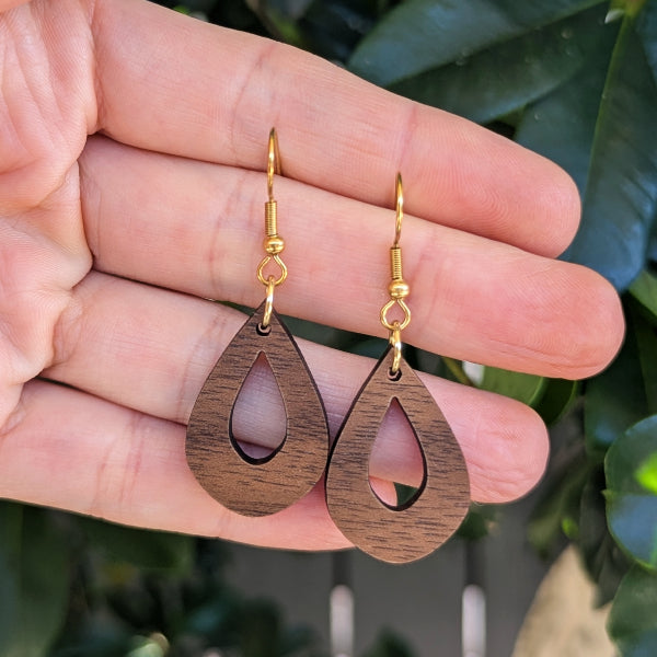 A pair of wooden teardrop dangle earrings with gold fishhook wires, held in a person's hand against a background of green leaves.
