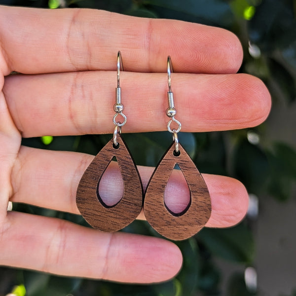 A pair of wooden teardrop dangle earrings with silver fishhook wires, held in a person's hand against a blurred background of green foliage.